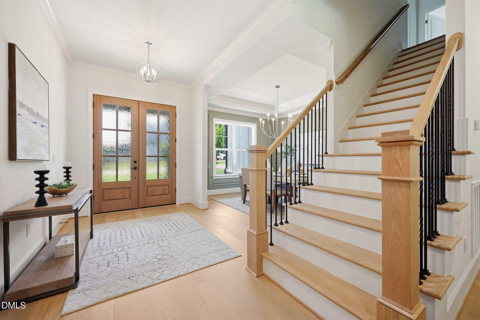 A bright, modern entryway with light wood floors, a wooden staircase with black spindles, double front doors with glass panels, a console table, and a dining room visible through an open doorway.