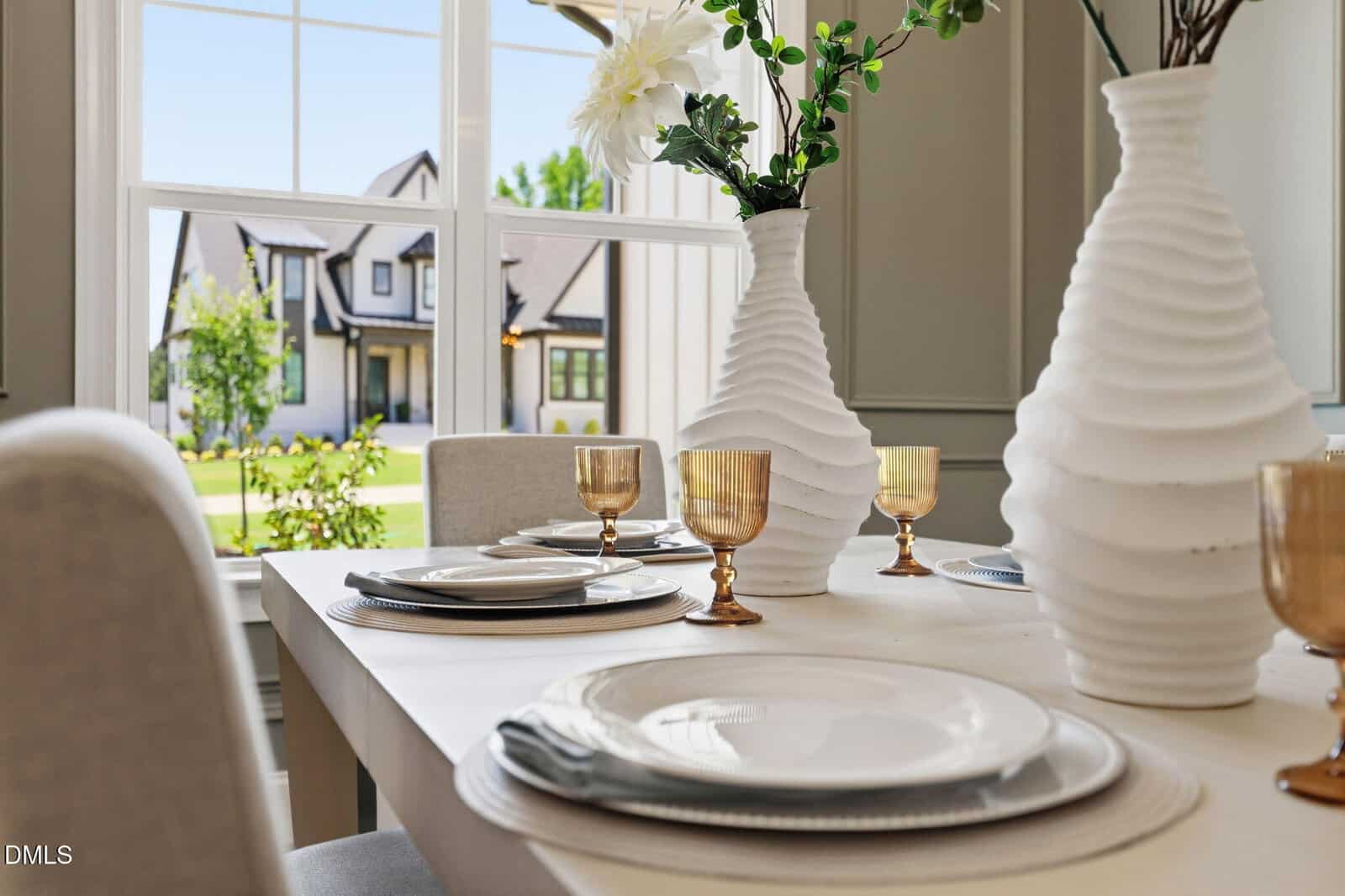 A dining table set with white plates, gold-rimmed glasses, and two white vases holding flowers, positioned by a window overlooking a suburban house and green lawn.