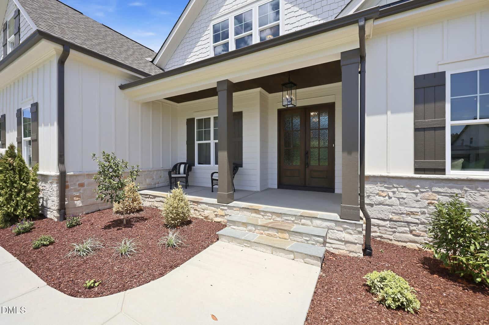 A modern house with white siding, stone accents, and a covered front porch. Two black chairs sit on the porch, and landscaped bushes are planted in mulched beds along a concrete walkway.