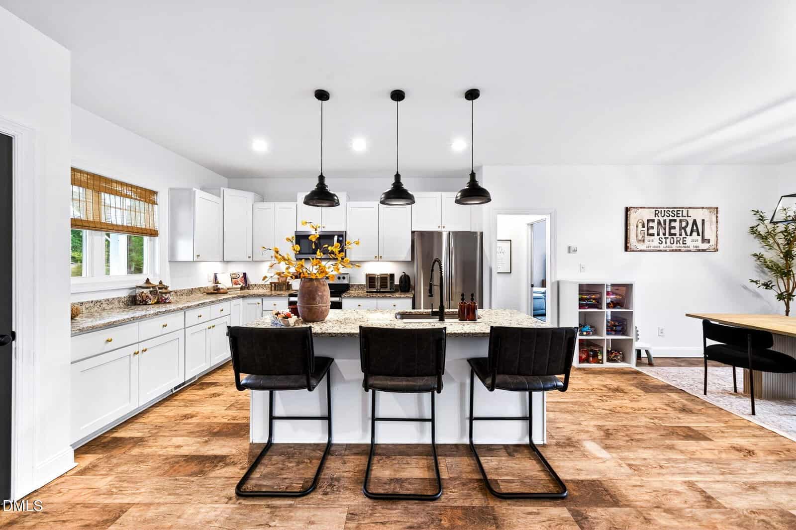 Modern kitchen with white cabinets, stainless steel appliances, a large island with granite countertop, black pendant lights, and four black chairs. Wooden floors and decor add warmth; dining and living areas are visible.