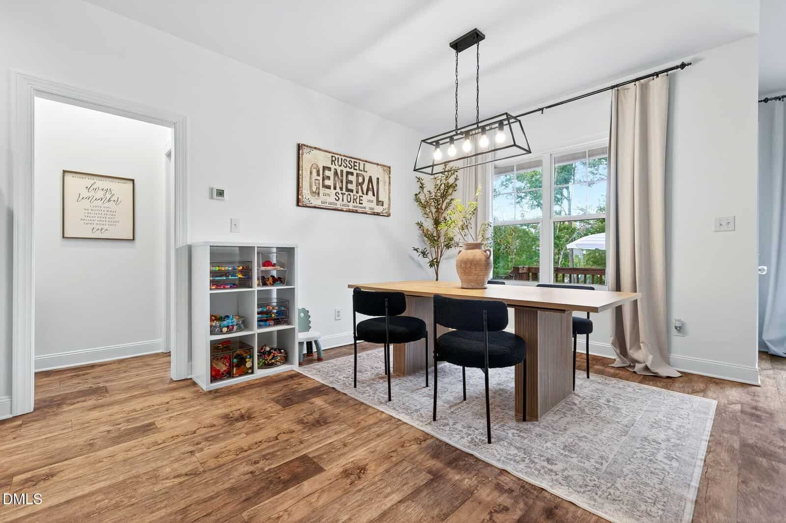 A dining area with a wooden table, four black chairs, a geometric light fixture, a window with curtains, a toy shelf, and wall art reading “Russell General Store.” Rug covers light hardwood floor.