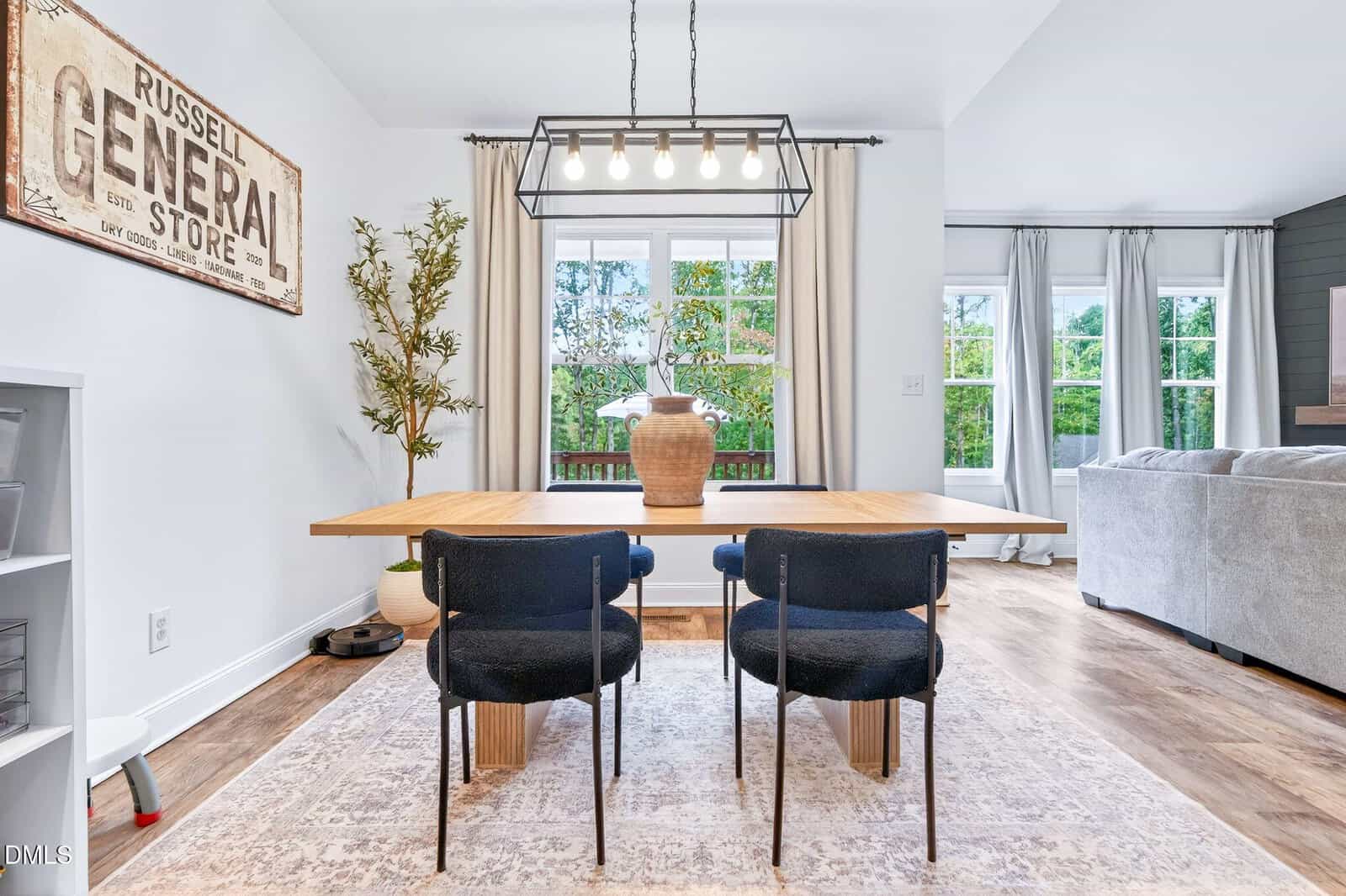 A modern dining area with a wooden table, four dark chairs, a large light fixture, a potted plant, and a “General Store” sign on the wall. Large windows let in natural light and overlook greenery outside.