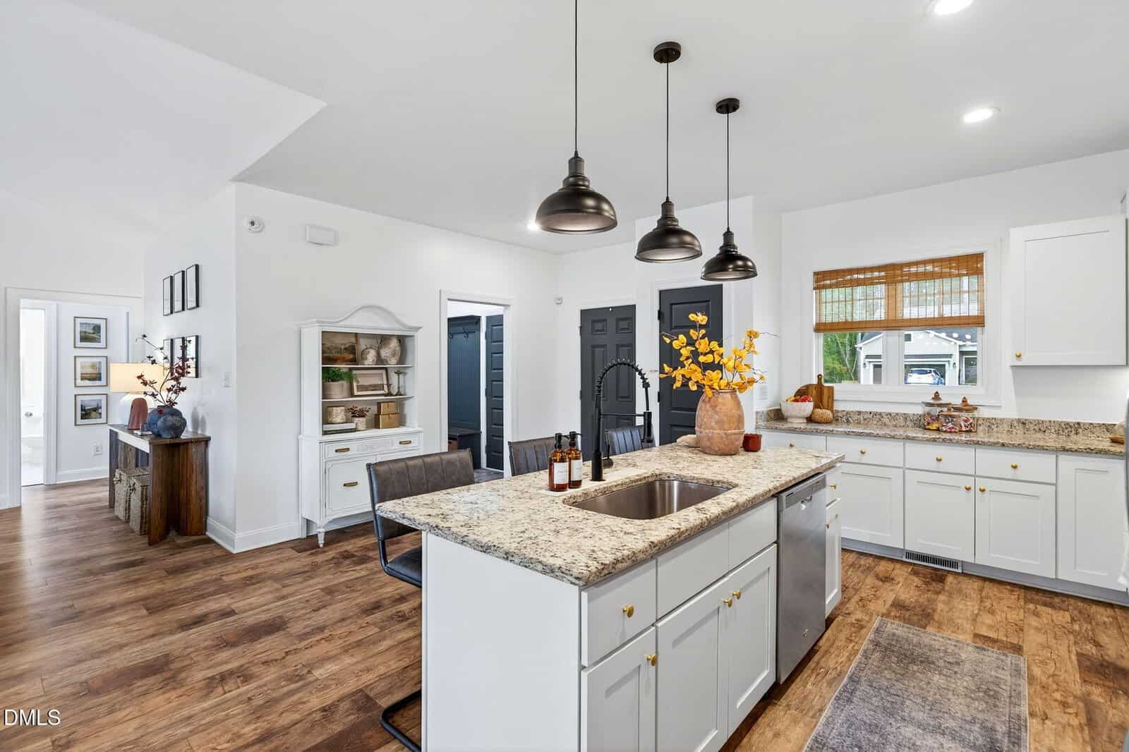 Modern kitchen with white cabinets, granite countertops, a central island with a sink, pendant lights, wood flooring, and decorative accents. A window with a bamboo shade overlooks a green yard, and adjacent rooms are visible.