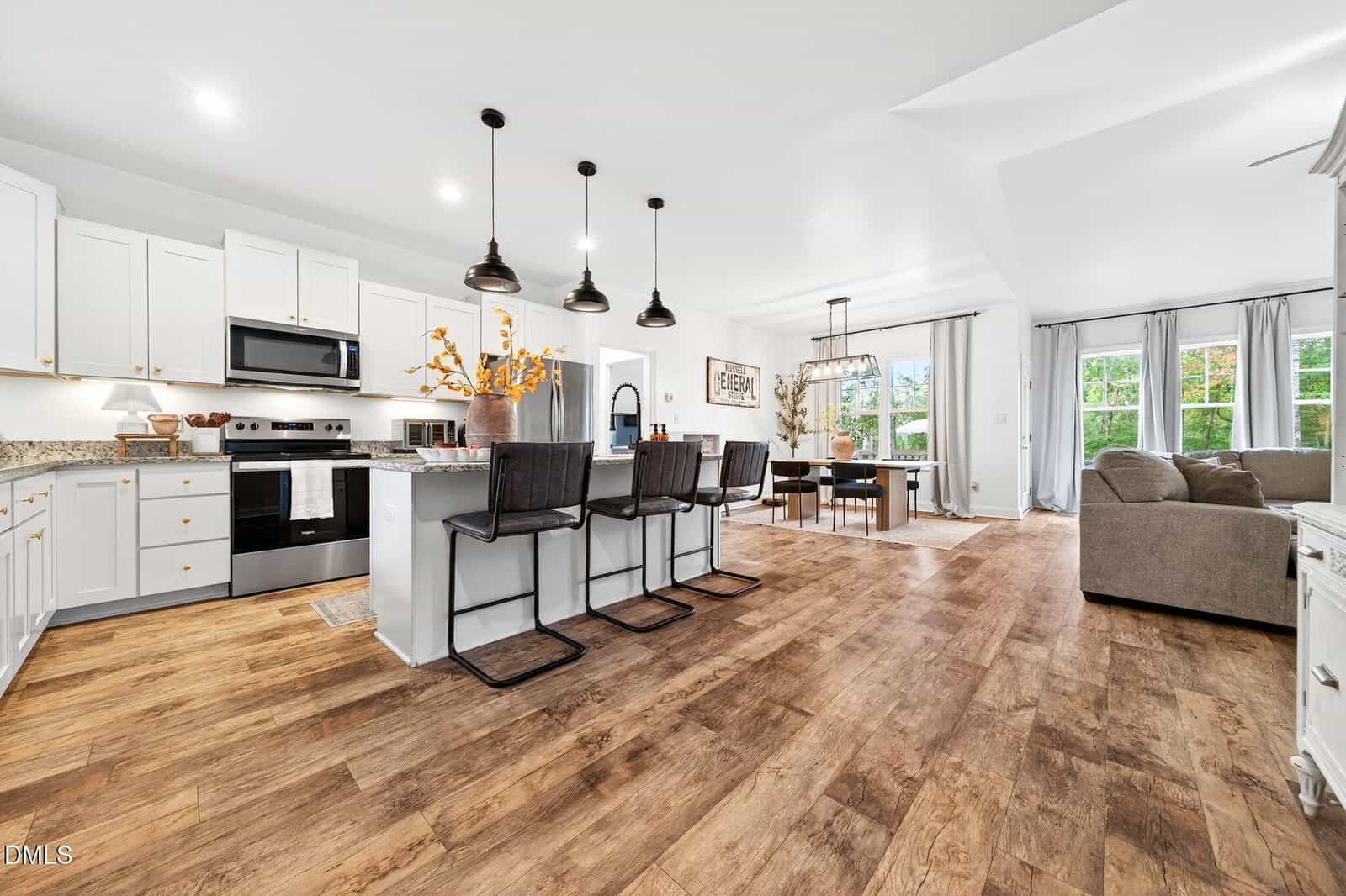 Open-concept kitchen and living area with white cabinets, granite countertops, black appliances, a large island with four dark barstools, dining table, gray sofa, wood floors, and large windows letting in natural light.
