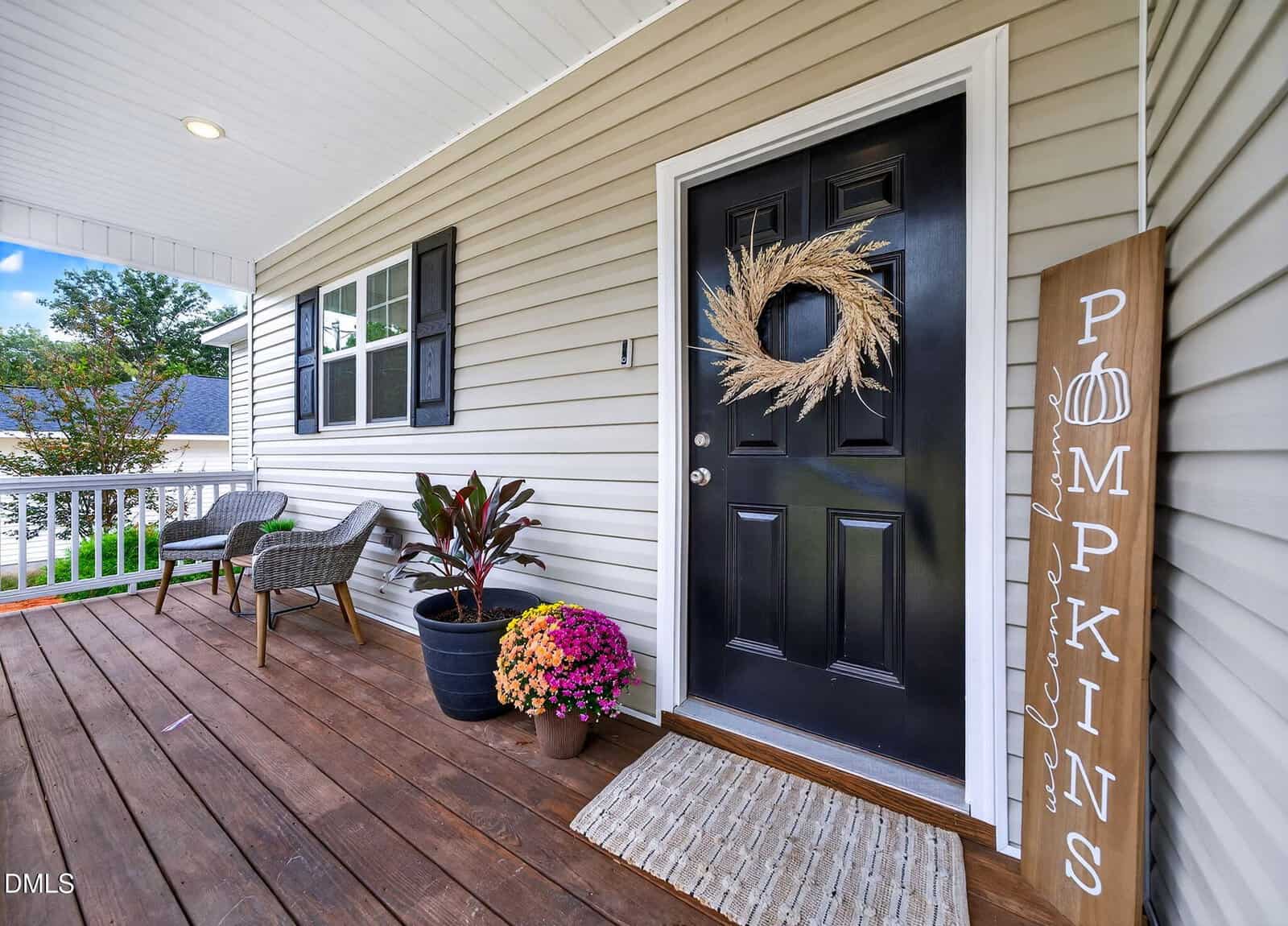 A cozy porch with wooden flooring, a black front door decorated with a wreath, potted plants, two chairs with a small table, and a vertical sign reading "PUMPKINS" beside a doormat.
