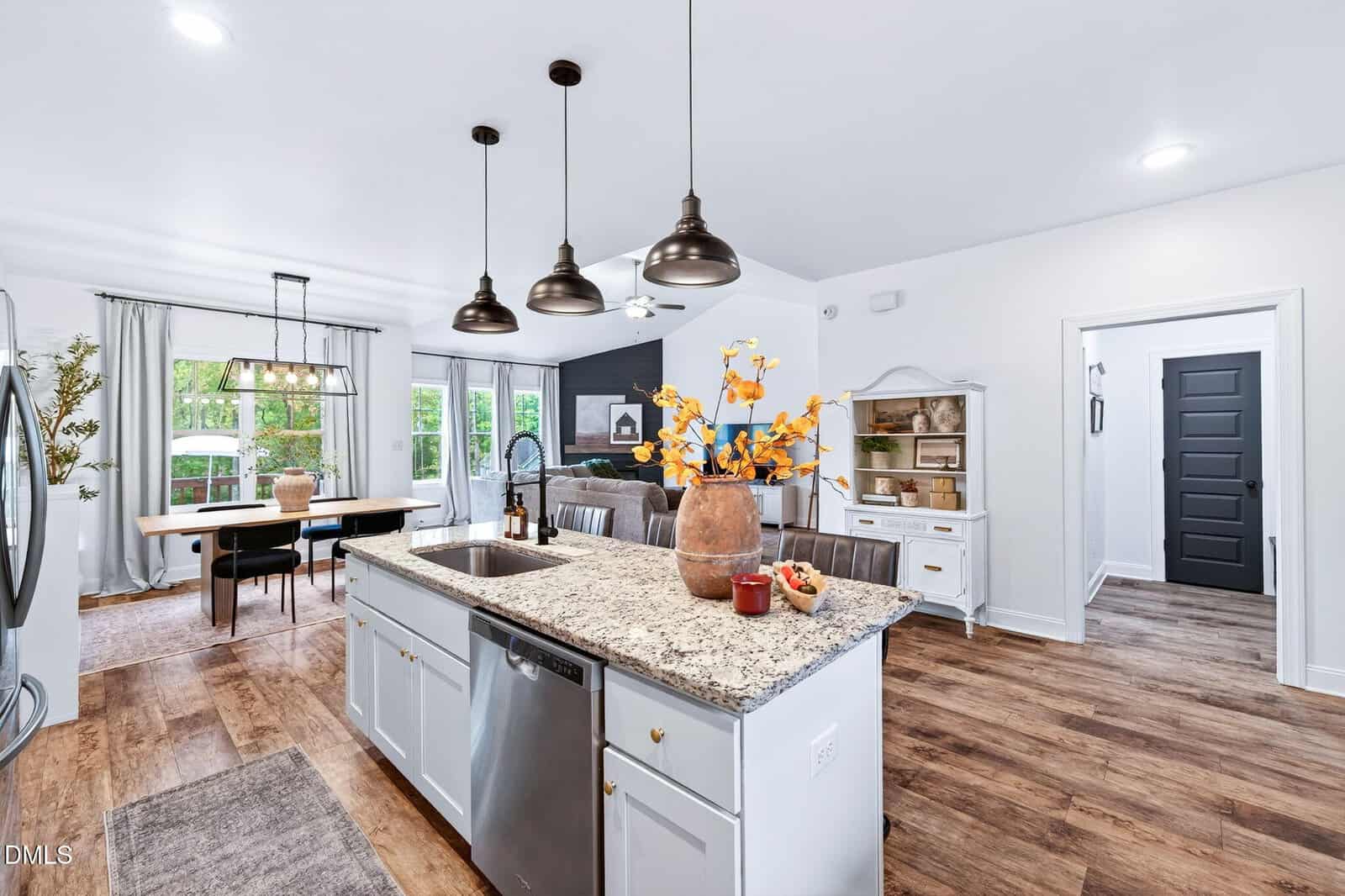 Bright modern kitchen with a granite island, farmhouse sink, white cabinets, and pendant lights. Dining area with large windows and wooden table in the background. Hardwood floors throughout the open-concept space.