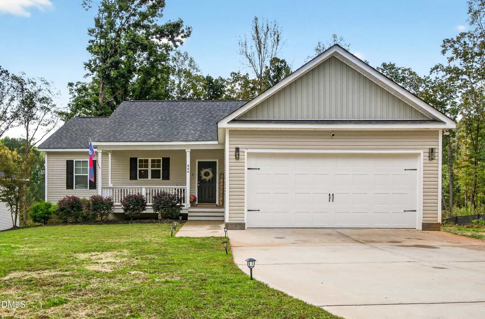 Charming single-story house in Louisburg with a gable roof, light gray siding, large white garage door, small front porch with railing, shrubs, and a green lawn at 644 Shawnee Drive, set against trees and a clear sky.