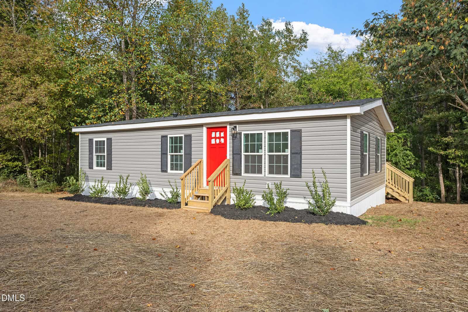 A single-story gray manufactured home with white trim and a red front door sits on a cleared lot at 3077 Rock Spring Church Road, Creedmoor, NC 27522 10127189, featuring mulch, small shrubs in front, and a backdrop of trees and greenery.