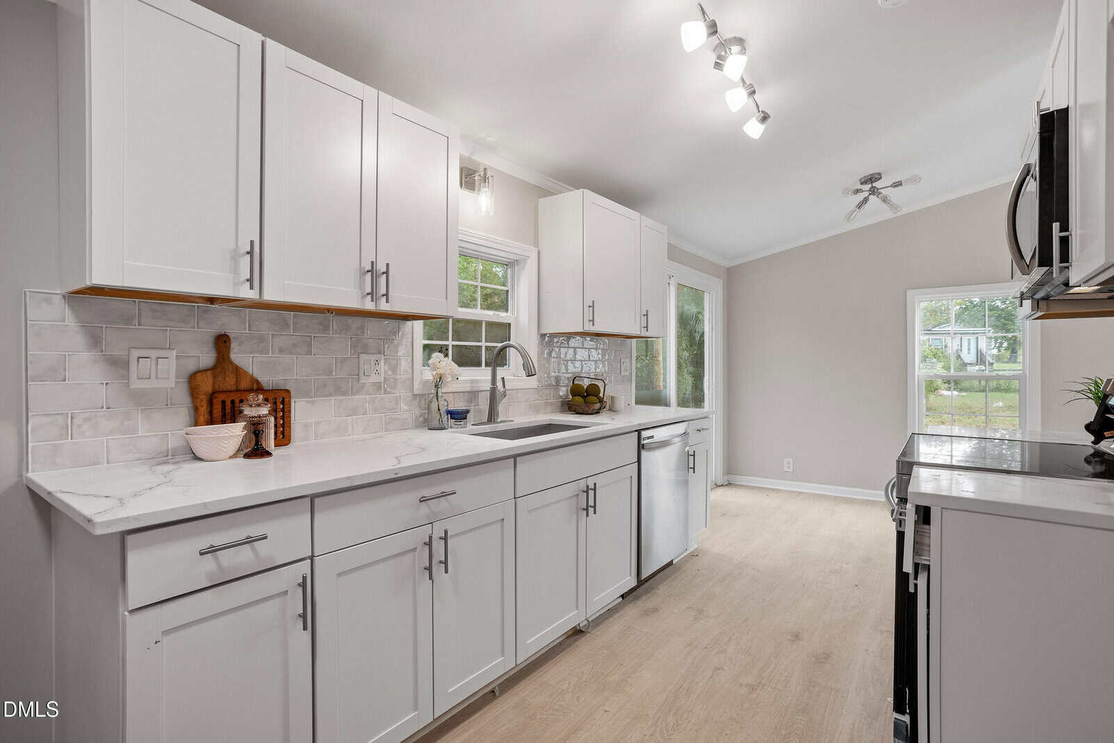 A modern kitchen with white cabinets, marble countertops, and stainless steel appliances. Light wood flooring and neutral walls brighten the room. Natural light comes in from windows and a glass door at the far end.