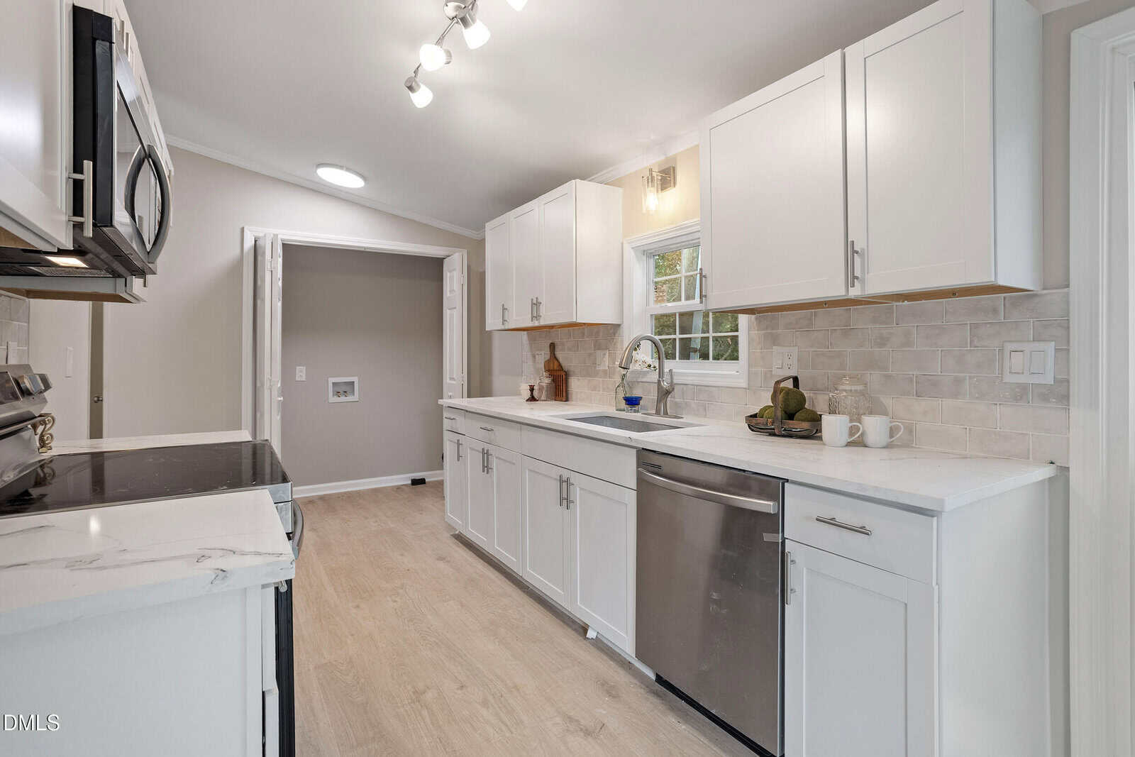A modern kitchen with white cabinets, stainless steel appliances, light wood flooring, beige tile backsplash, and a window above the sink. The room is well-lit with ceiling lights.
