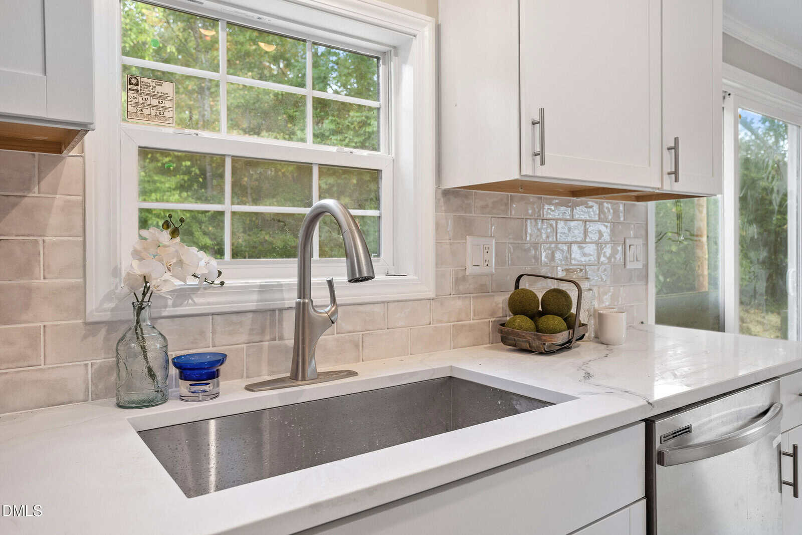 A modern kitchen sink with a stainless steel faucet is set in a white countertop. A vase with white flowers, a blue container, and a tray with green decorative spheres are next to the sink. A window is above the sink.