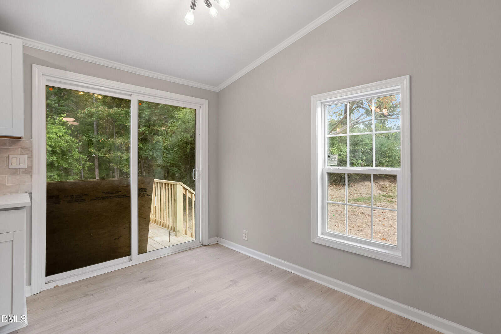 A room with light-colored wood flooring, beige walls, a white-framed window, and a glass sliding door leading to a deck outside. Trees and greenery are visible through the window and door.