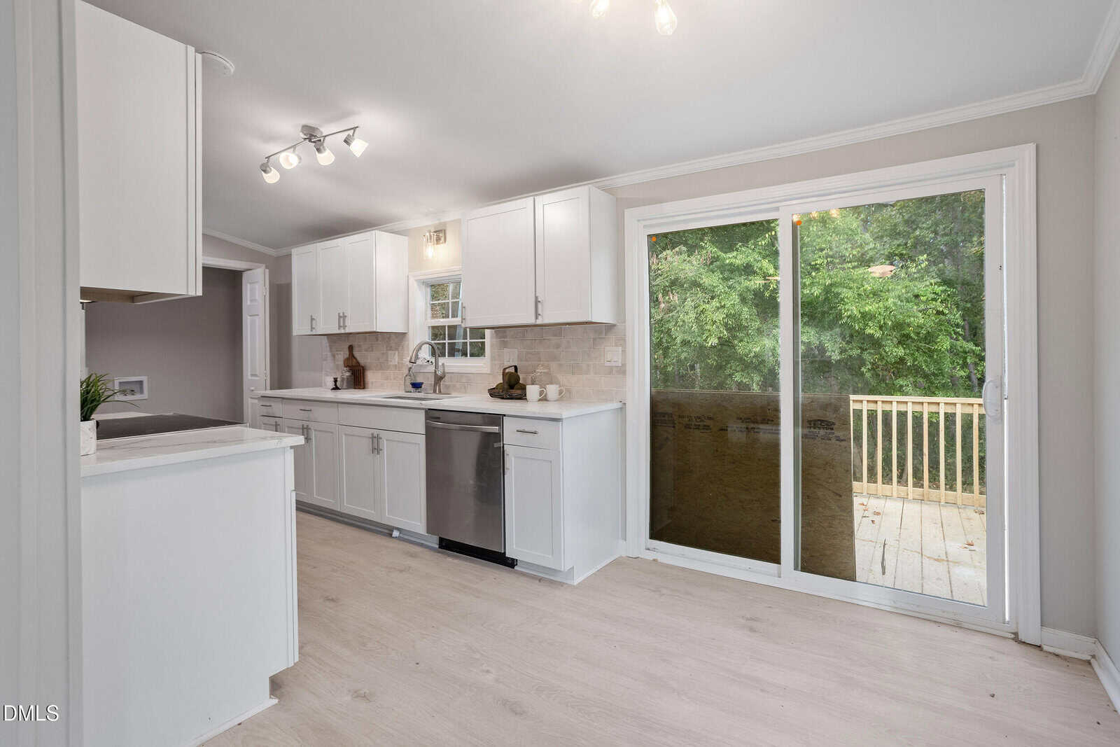 A modern kitchen with white cabinets, light wood flooring, stainless steel appliances, and a sliding glass door leading to a wooden deck with a view of green trees outside.
