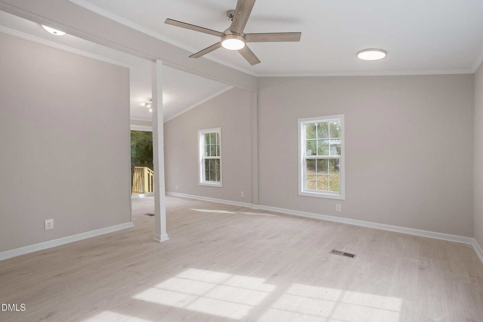 A spacious, empty room with light-colored wood flooring, beige walls, two windows, a ceiling fan, and recessed lighting. An open doorway leads to a deck outside. Natural light fills the space.