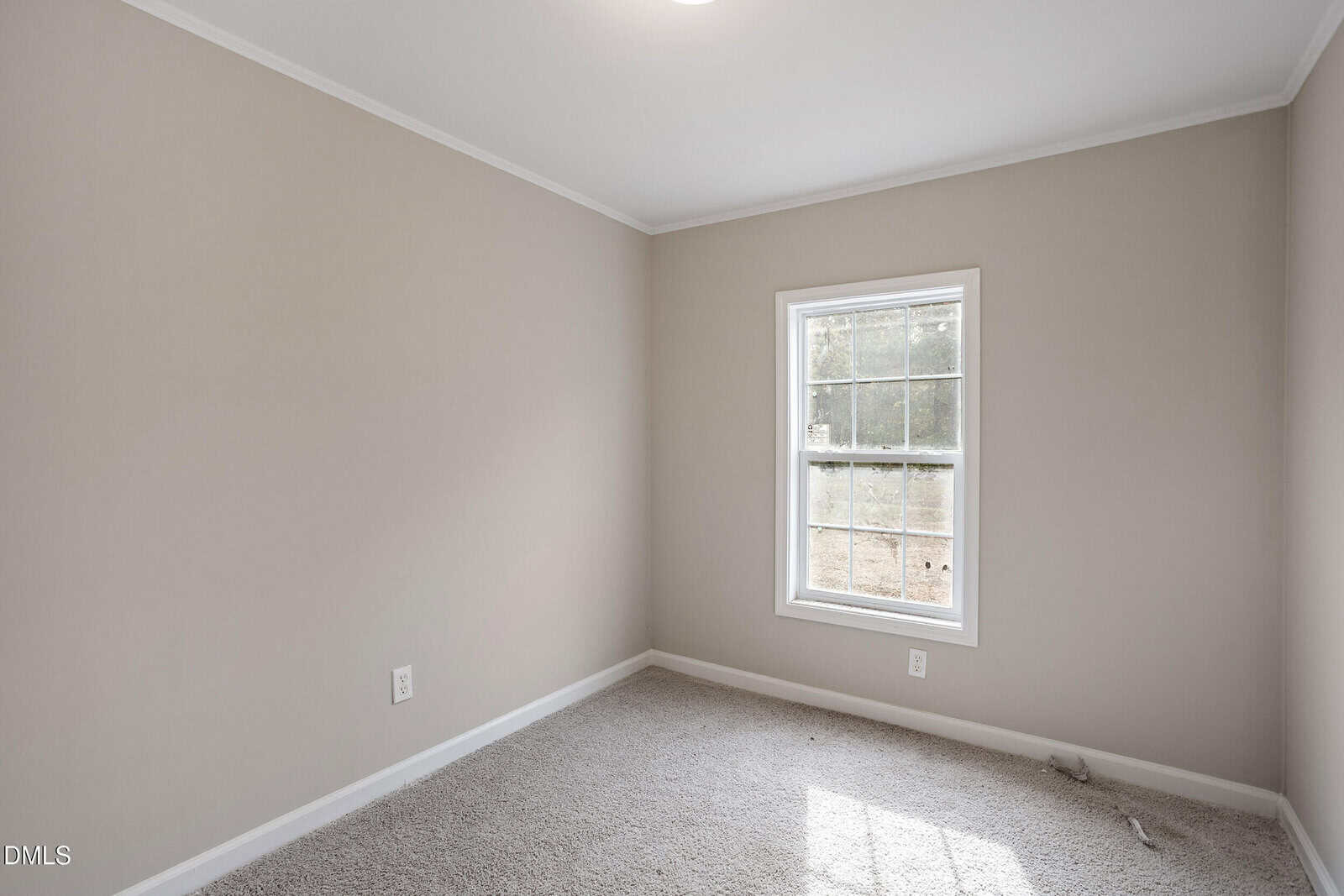 A small, empty room with beige walls, white trim, a single window letting in daylight, and light-colored carpet flooring. The room has a clean and minimal appearance.