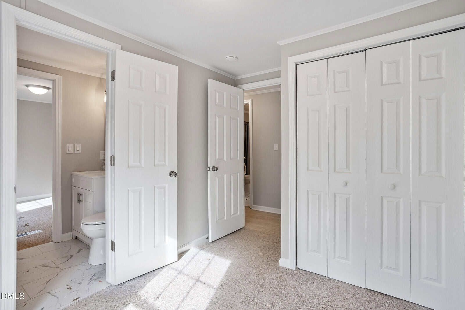 A small, carpeted bedroom with light gray walls, white trim, double closet doors, and an open door leading to a bathroom with a toilet and vanity. Sunlight shines in through an unseen window.