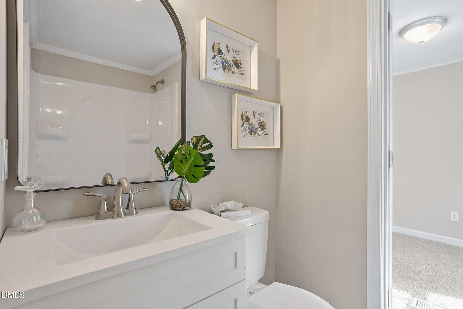 A modern bathroom with a white sink and faucet, a mirror above the sink, a toilet, two framed floral artworks on the wall, and a decorative plant on the counter. A shower with a curtain is partially visible.