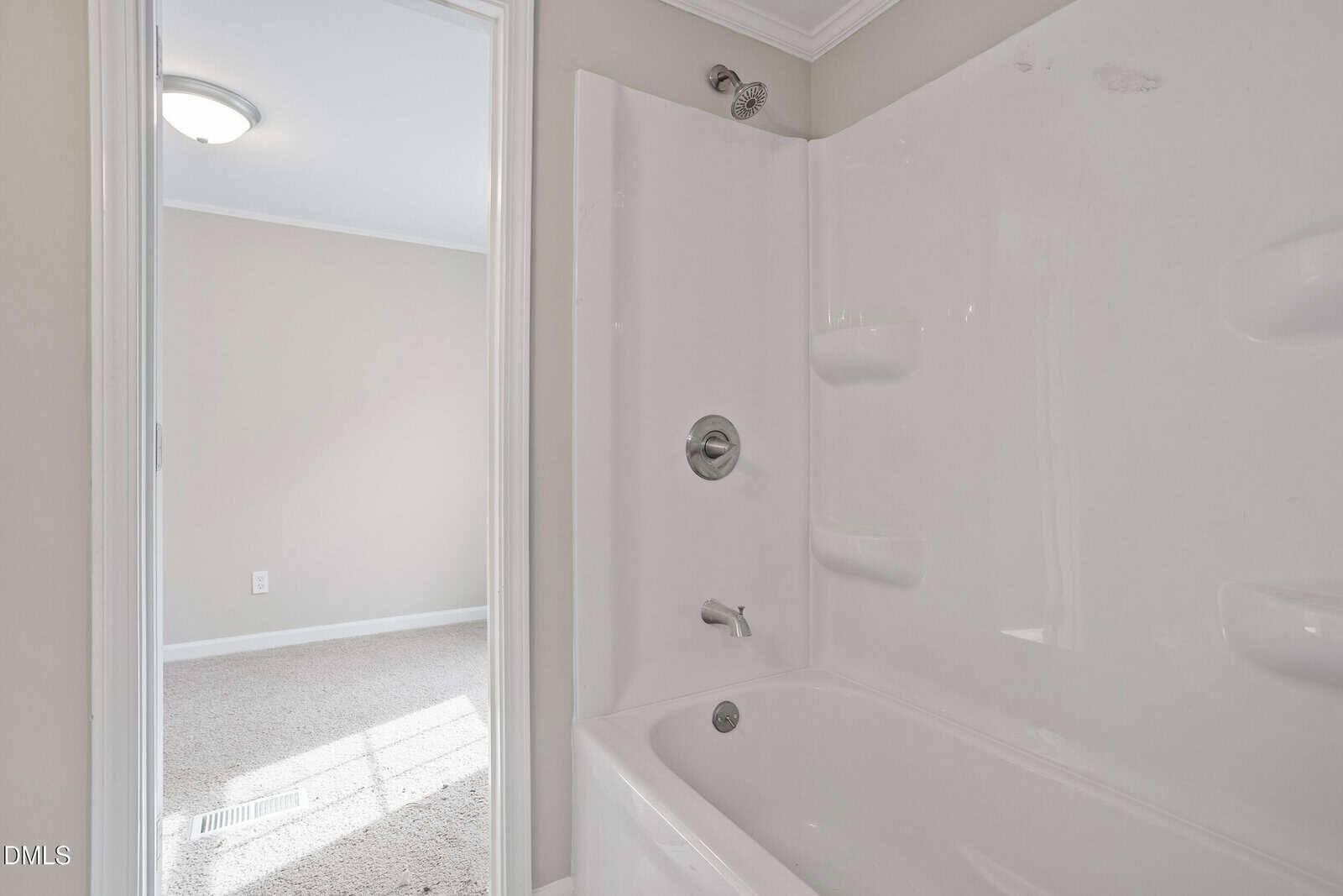 White bathtub and shower with built-in shelves in a bathroom; view through open door shows a carpeted room with beige walls, white trim, and a ceiling light.