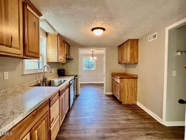 A kitchen with wood cabinets, marble-patterned countertops, stainless steel sink, and dark wood flooring. A window and light fixture are visible at the far end, with an adjacent open doorway on the right.