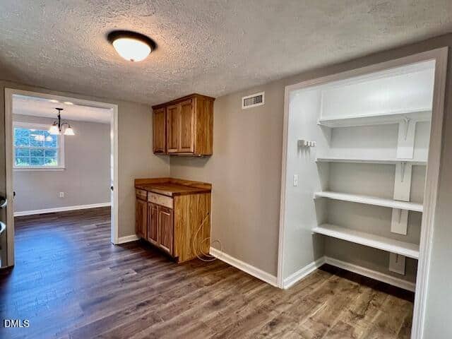 A pantry with white shelves is adjacent to a small space with wood cabinets and countertop; both rooms have wood-look flooring and neutral-colored walls. A dining area with windows is visible in the background.