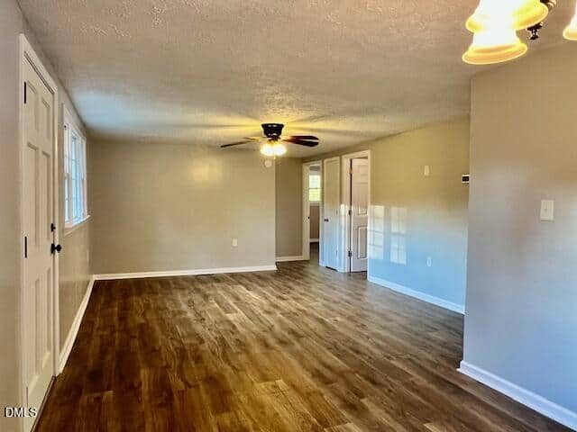 A spacious living area with light beige walls, wood-look flooring, a ceiling fan with lights, and multiple doors and windows. A chandelier hangs in the foreground on the right. The space appears empty and well-lit.
