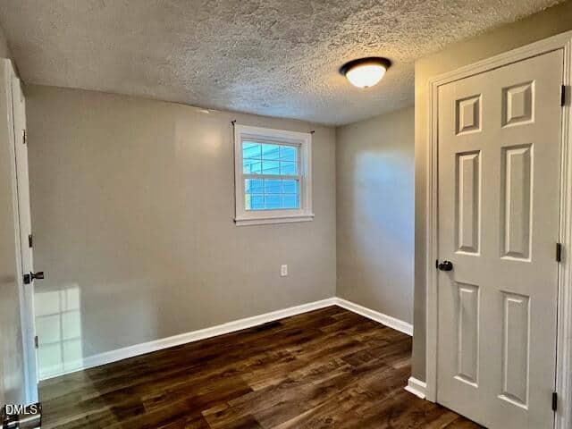 Small, empty room with beige walls, white trim, and dark wood flooring. There is a single window, a closed closet door, and a ceiling light fixture.