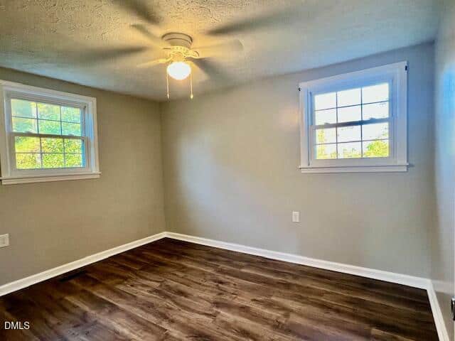 Empty room with light gray walls, dark wood flooring, two windows with white trim, and a ceiling fan with a light.