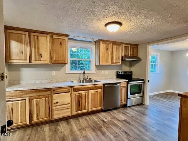 A kitchen with wooden cabinets, marble-look countertops, a stainless steel stove and dishwasher, a sink under a window, and wood-style flooring. An adjoining dining area with windows is visible through an open doorway.