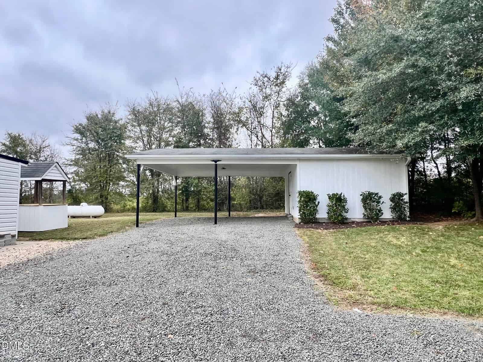 A gravel driveway leads to a white, open carport with a flat roof supported by black posts. A small lawn and shrubs are on the right, and trees form the background. A white shed and propane tank are to the left.
