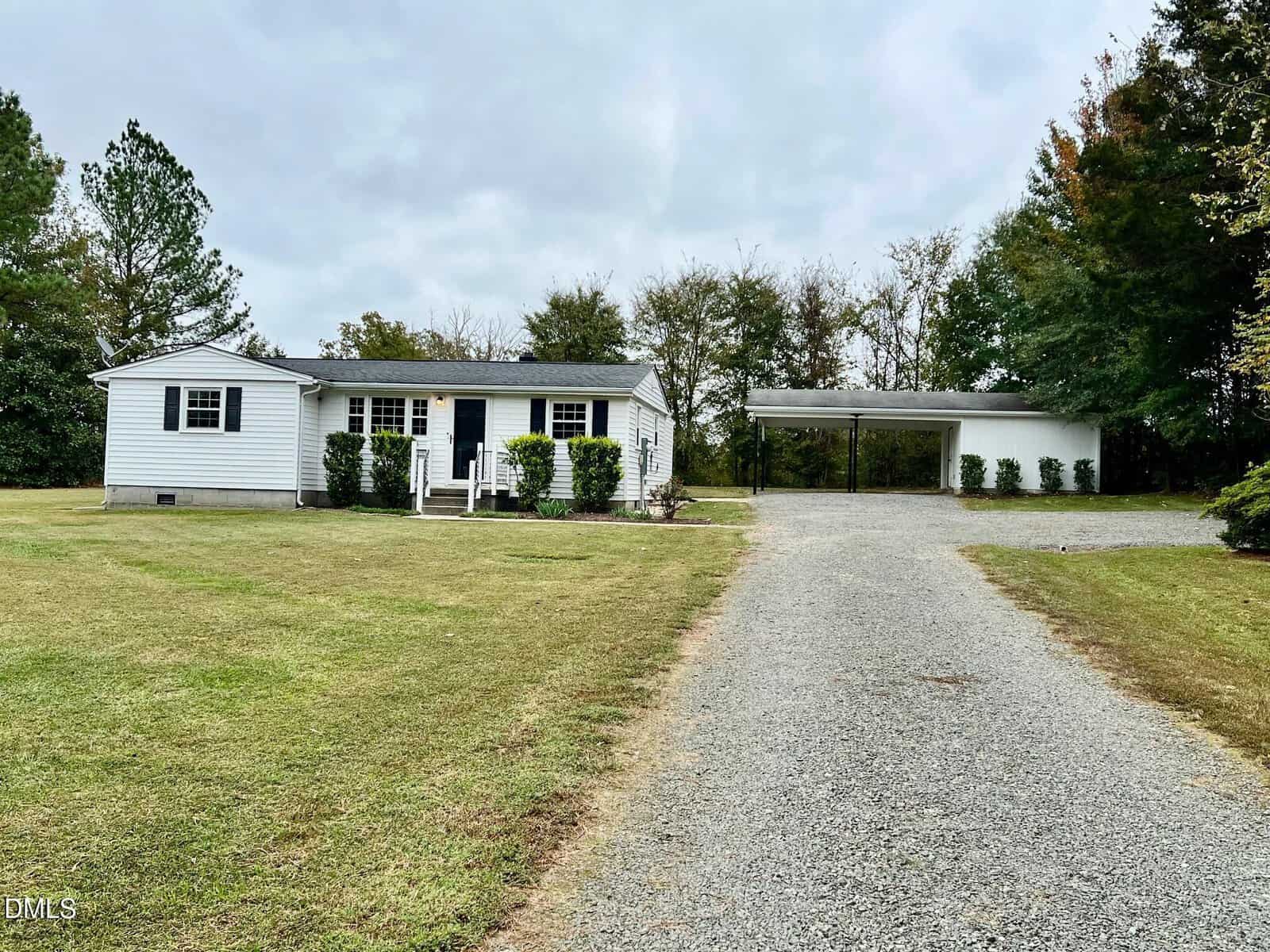A single-story white house with black shutters sits on a large grassy lot at 9590 Amis Chapel Road. A gravel driveway leads to a detached carport, and trees line the background under a cloudy sky.
