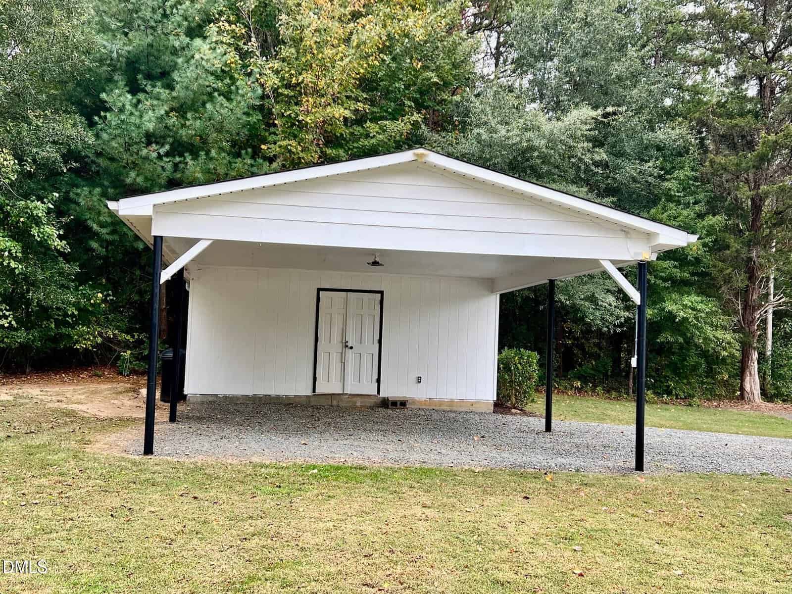 A white, single-car carport with a gable roof and black support posts stands on a gravel area in front of a wooded background. Double doors are centered on the back wall. Grass covers the foreground.