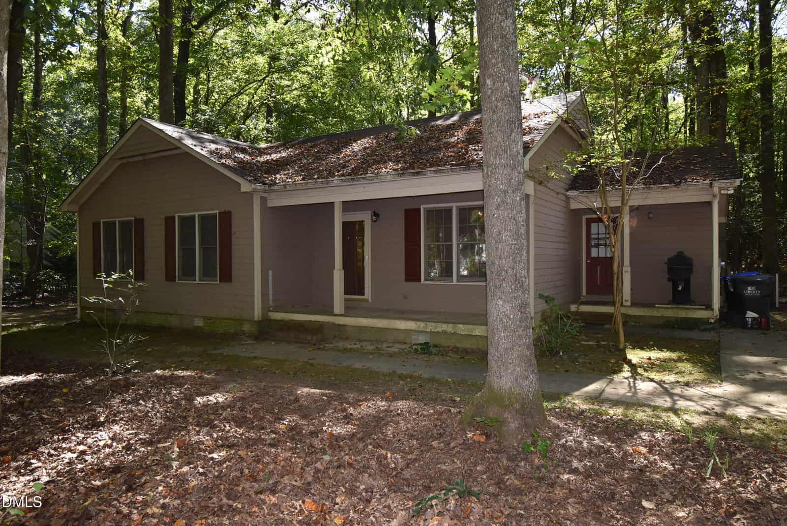 Nestled in a wooded area of Youngsville, this charming single-story house at 116 Thistle Drive features brown siding, a red front door, white trim, and a small front porch. The yard is shaded by trees, with leaves covering the ground and roof, offering a peaceful retreat surrounded by nature.