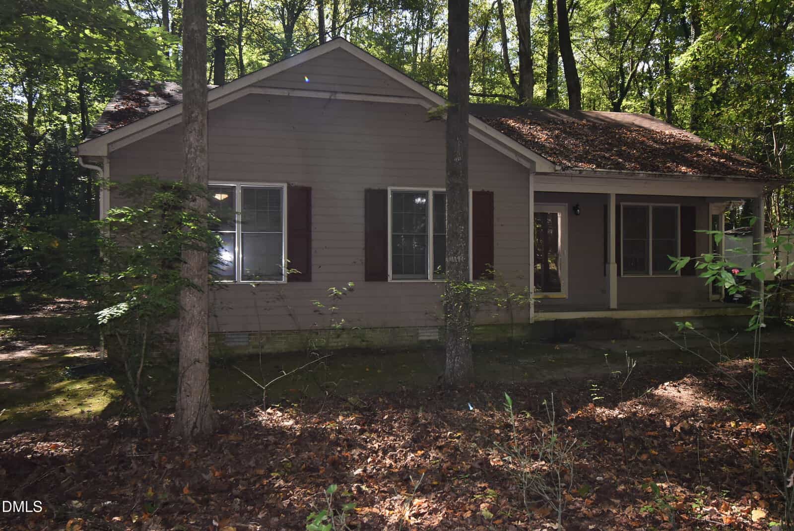 A single-story house with gray siding, brown shutters, and a brown roof is surrounded by trees. The yard is covered with fallen leaves and has patches of sunlight and shade. The house has multiple windows and sits in a wooded area.
