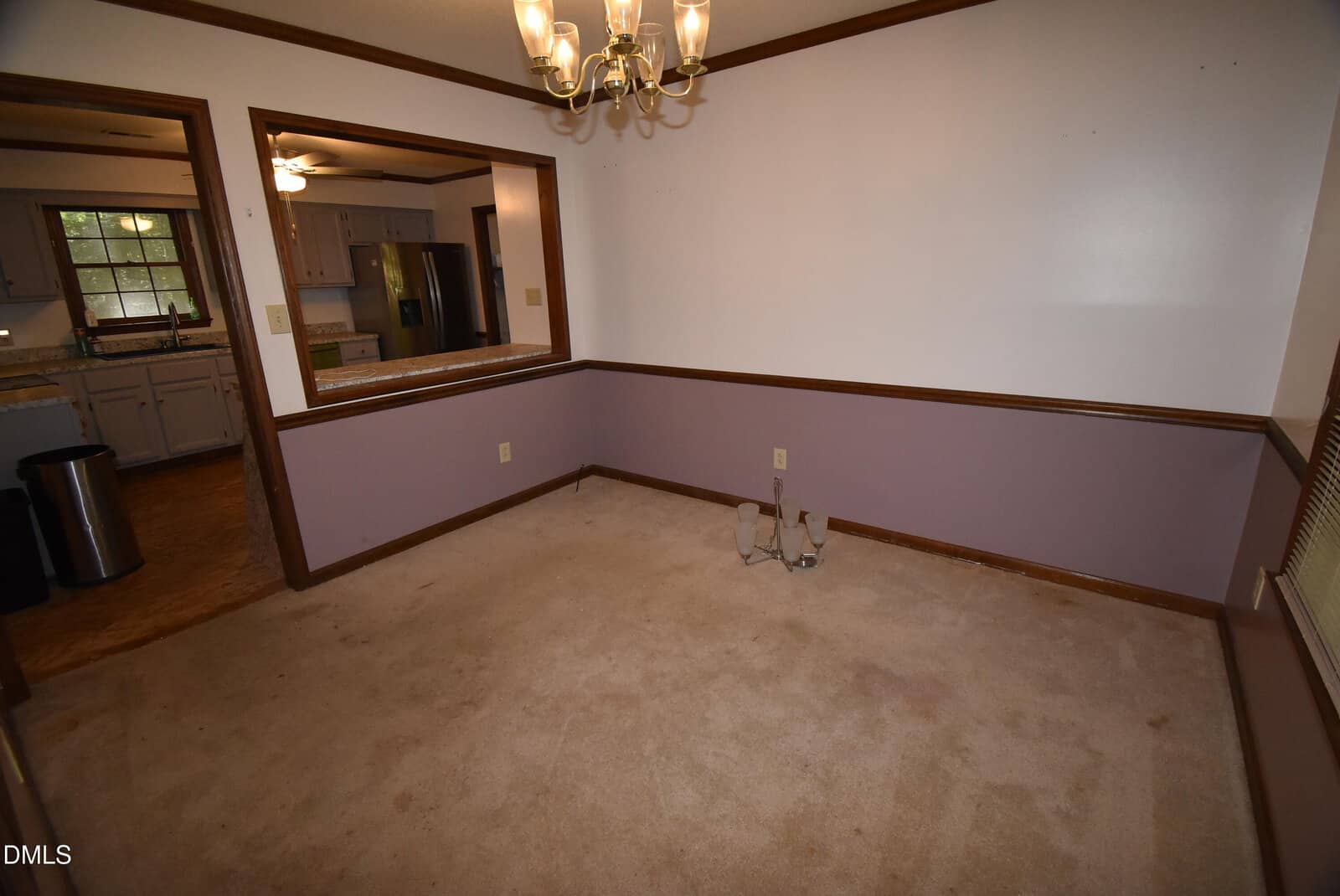Dining room with beige carpet, purple lower walls, white upper walls, wood trim, chandelier, electrical outlets, and two pipes on the floor. A pass-through window opens to a kitchen with visible cabinets, sink, and refrigerator.