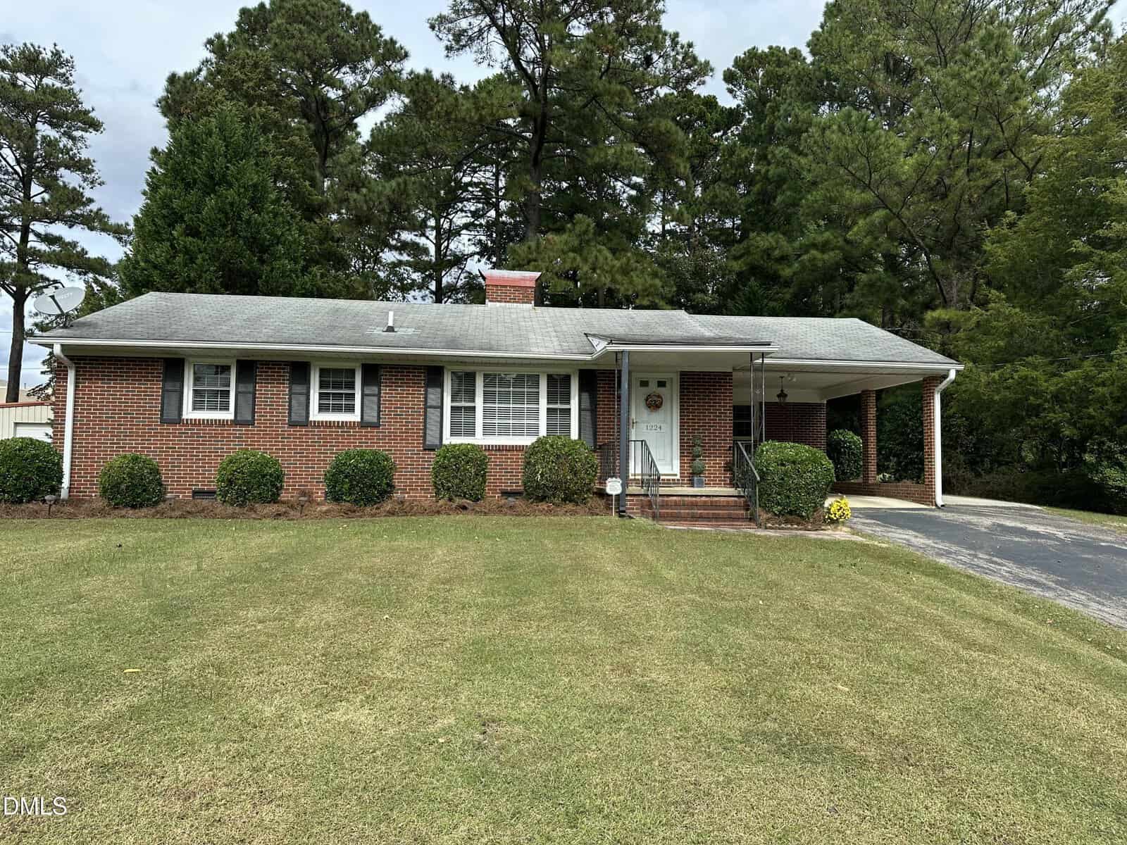 Charming single-story red brick house with white trim at 1224 Oakridge Avenue in Henderson. Features a front porch, carport on the right, green shrubs, a well-kept lawn, and tall trees in the background.