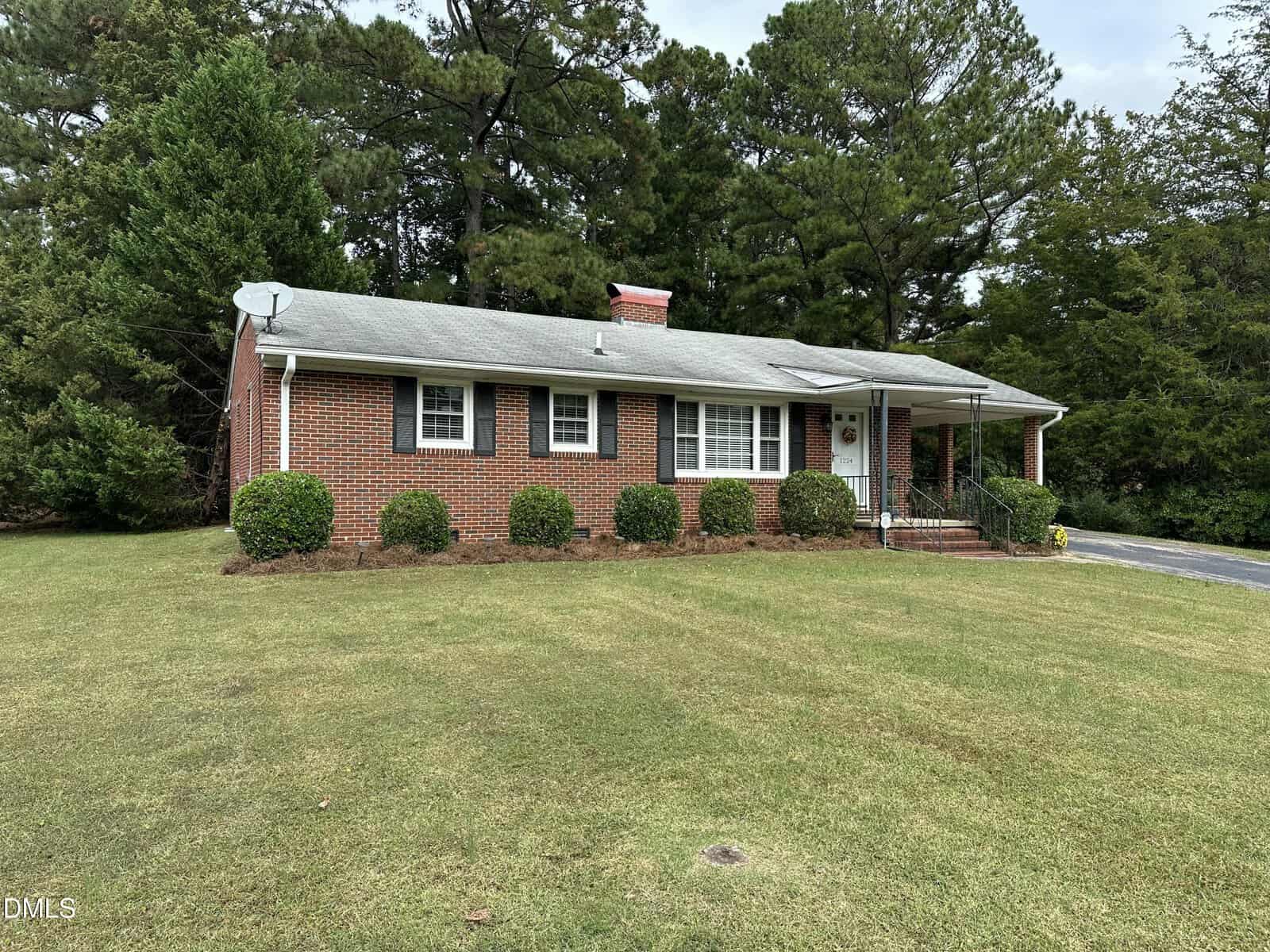 A single-story red brick house with black shutters, a gray roof, and a small covered front porch sits on a neatly mowed lawn, surrounded by trees. A driveway is visible on the right side.