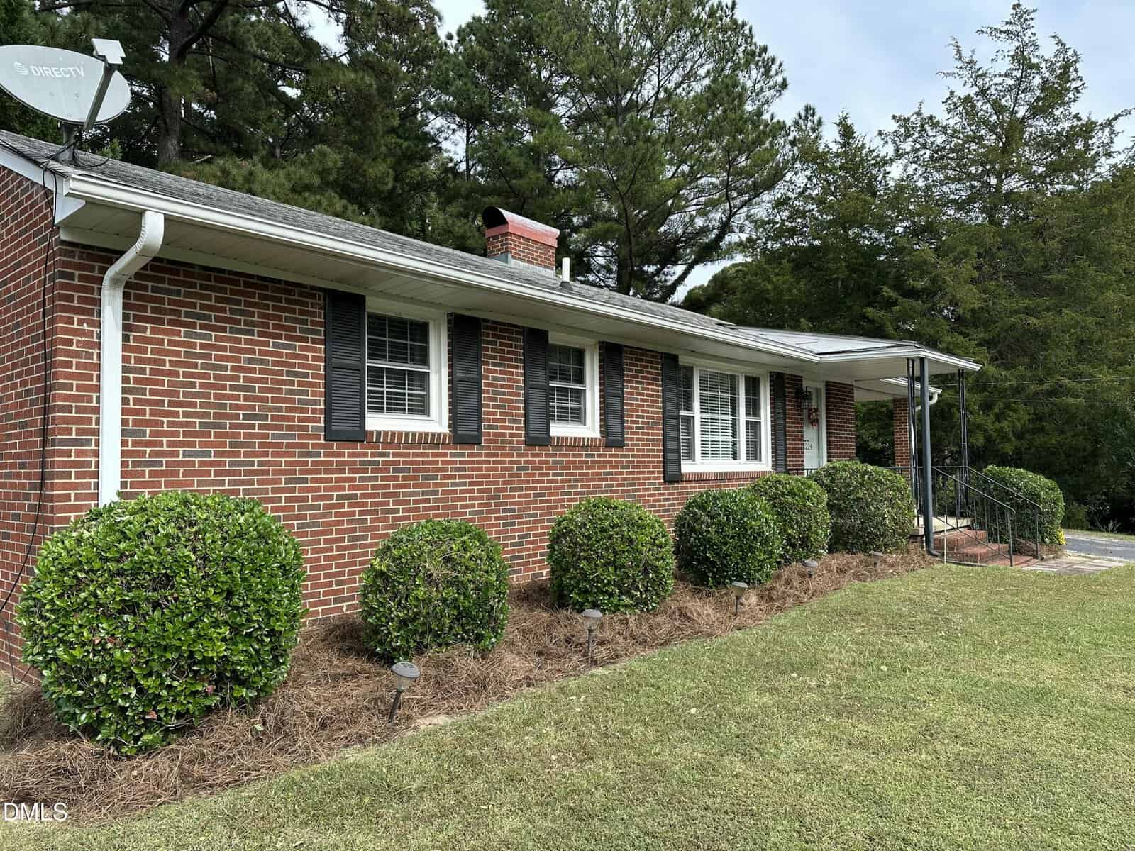 A one-story red brick house with black shutters, white trim, and a gray roof, surrounded by green bushes and pine straw mulch along the front, with trees in the background.