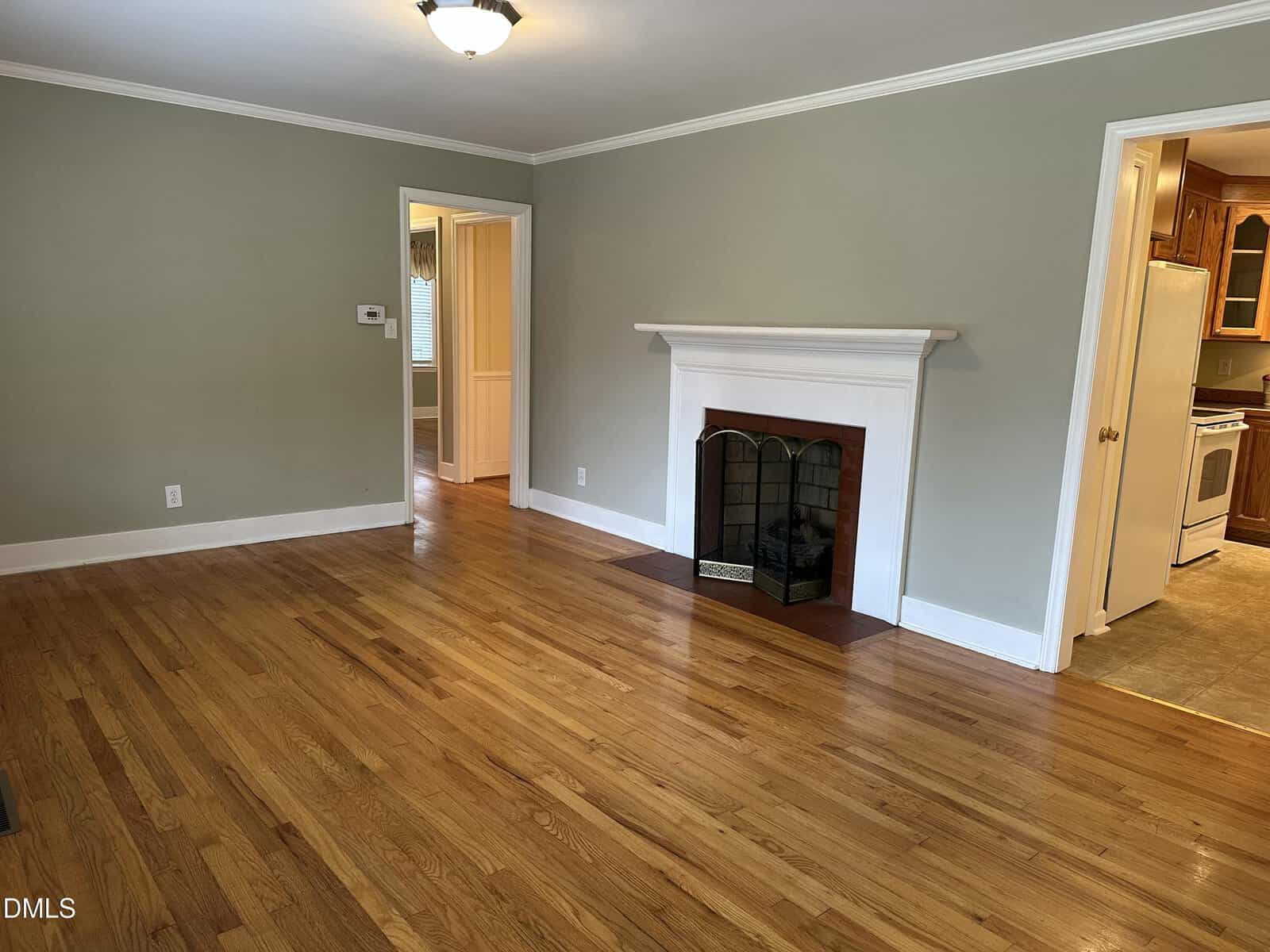 A bright living room with hardwood floors, a white fireplace with a gold screen, light gray walls, and white trim. Two doorways lead to adjacent rooms, one of which is a kitchen with wooden cabinets.