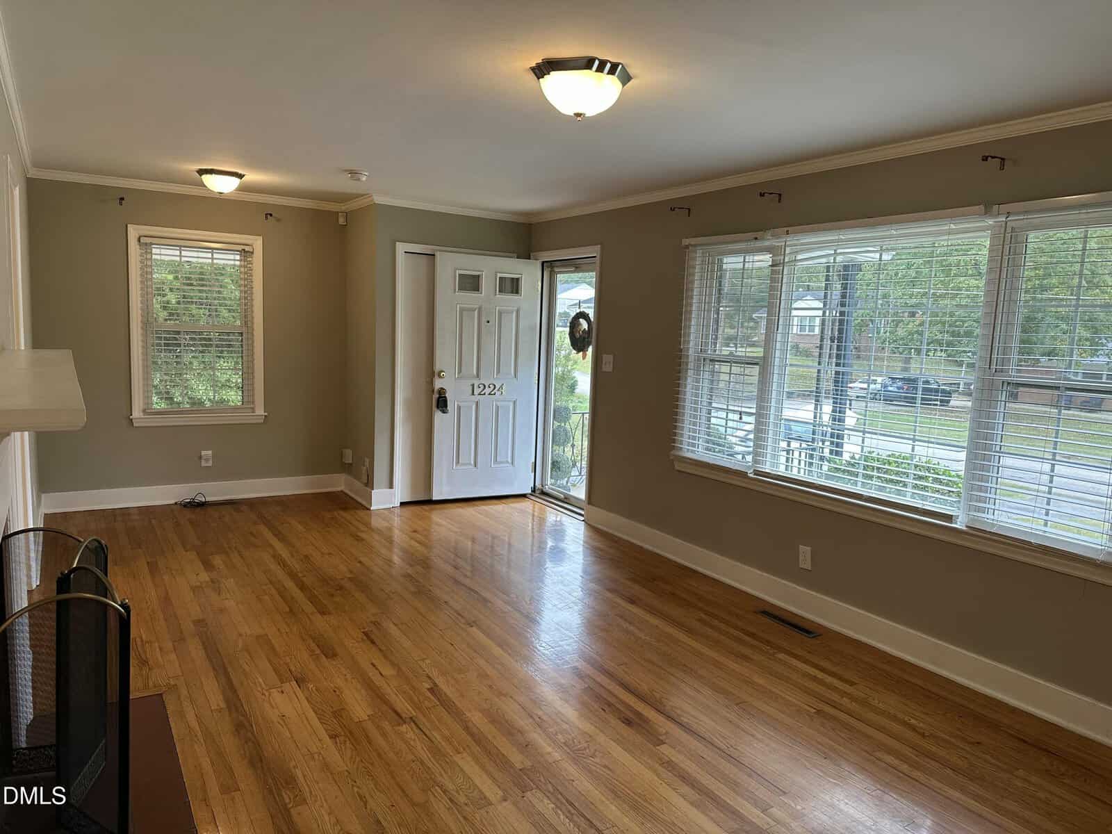 Bright living room with hardwood floors, large windows with blinds, a white front door with glass panels, beige walls, ceiling lights, and a fireplace partially visible on the left.