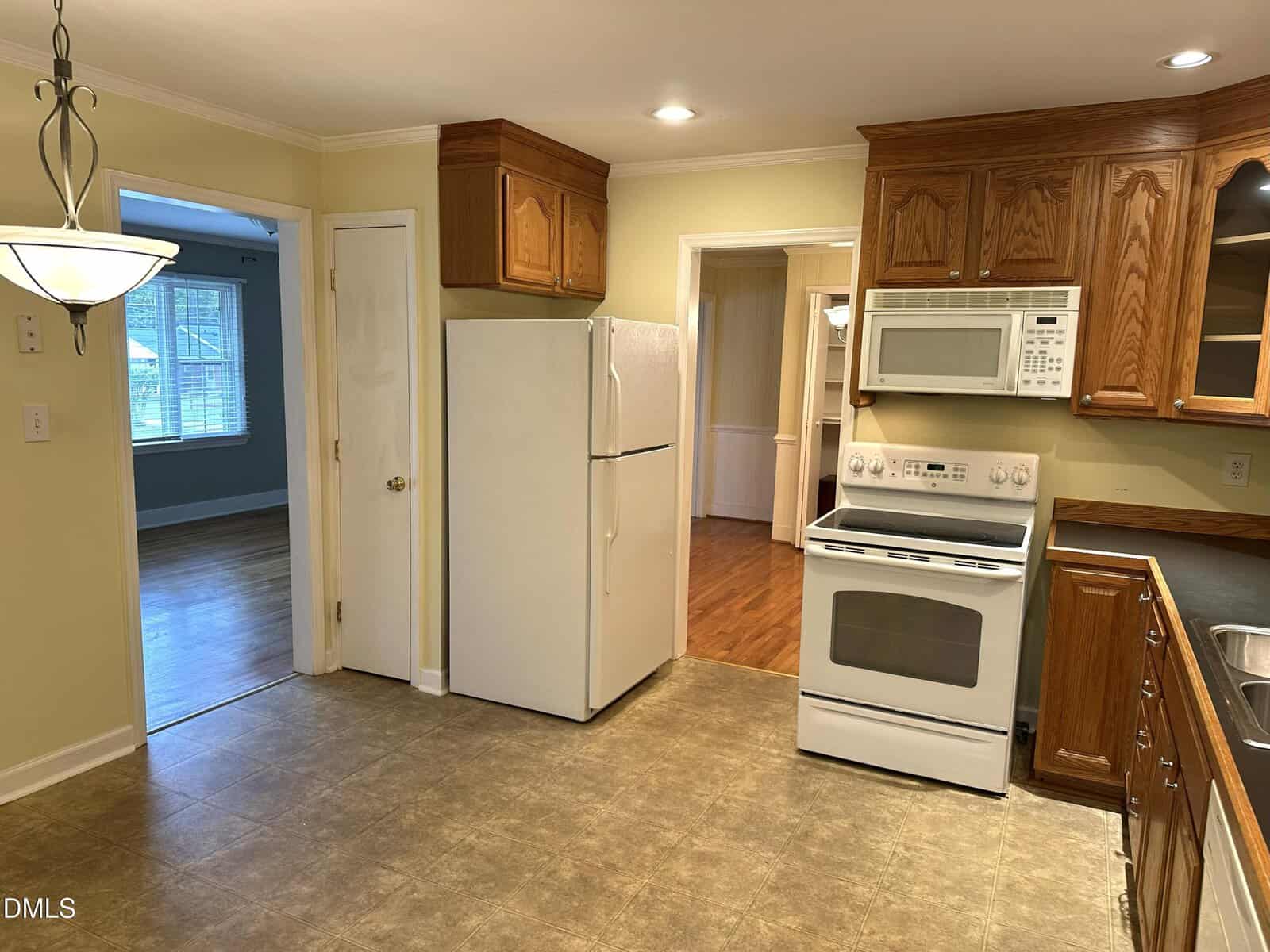 A kitchen with wooden cabinets, white appliances including a refrigerator, stove, and microwave, and tan tile flooring. An adjacent dining area and living room with hardwood floors are visible through open doorways.