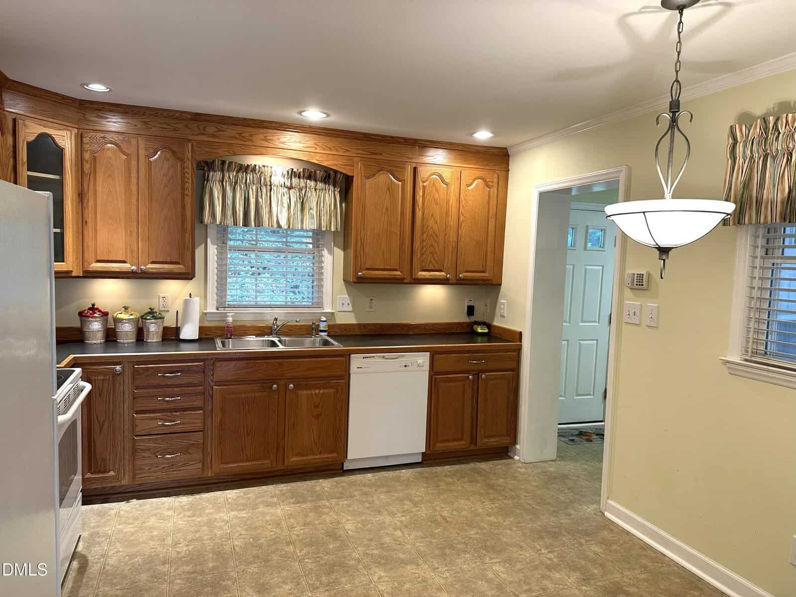 A kitchen with wooden cabinets, a white dishwasher, sink, and stove. There is a window with a valance above the sink, a hanging light fixture, and a door leading to another room. The floor is beige tile.