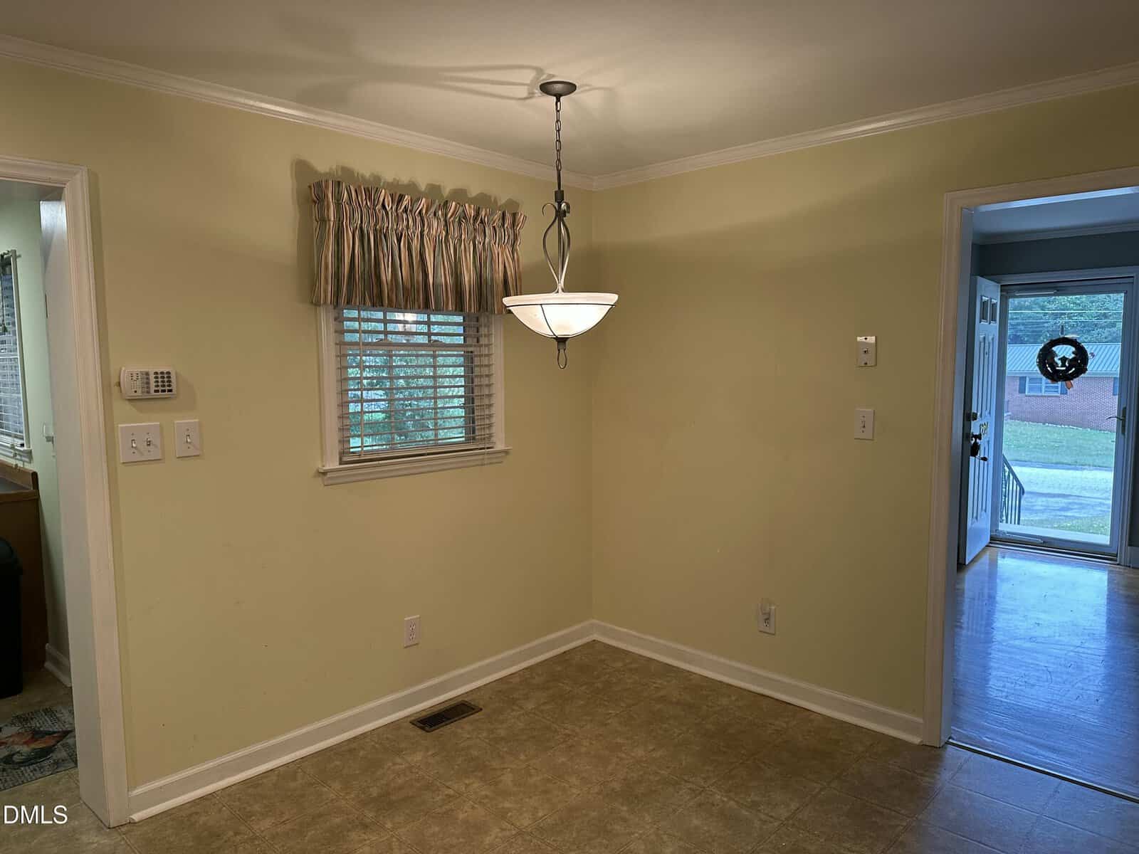 A small dining area with yellow walls, a window with valance, hanging light fixture, tiled floor, and view into a kitchen and hallway with front door.