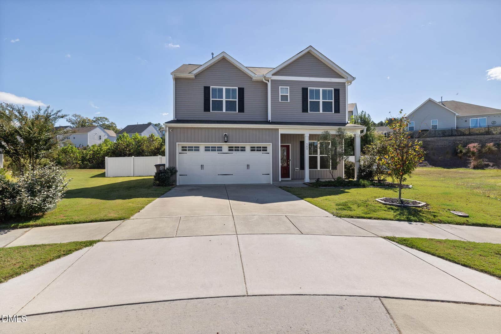 Two-story suburban house with gray siding, black shutters, a double garage, and a manicured lawn. The driveway is wide and bordered by grass, with a small tree in the yard and neighboring houses visible in the background.