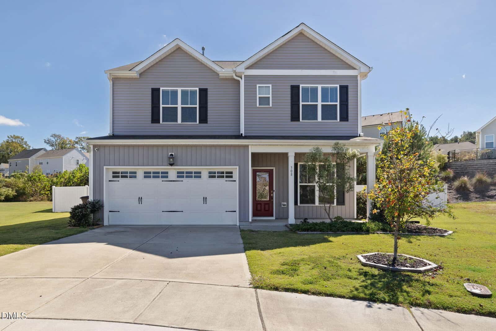 Two-story suburban house at 308 Colin Court in Wake Forest, featuring gray siding, white trim, black shutters, and a striking red front door. Attached two-car garage with white doors. Concrete driveway, small front lawn, and young trees in landscaped beds. Clear, sunny sky.