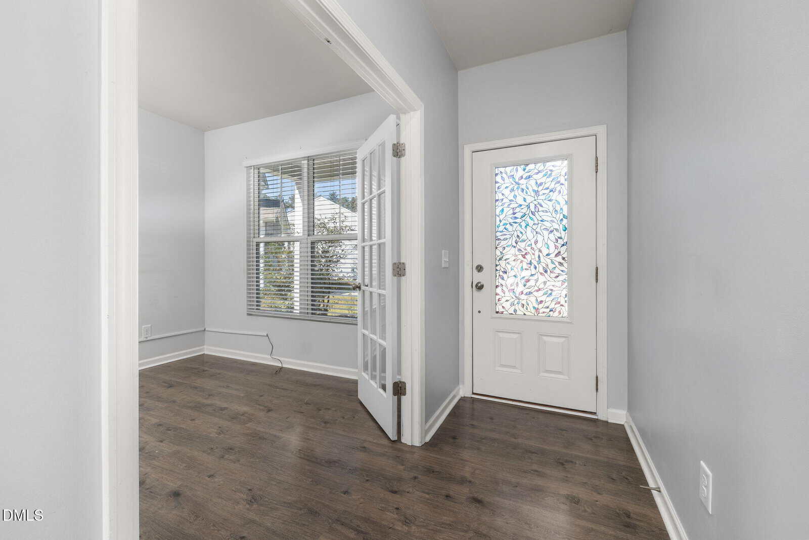 A small entryway with light gray walls, dark wood floors, a white door with a decorative frosted glass window, and French doors leading to a bright room with large windows.