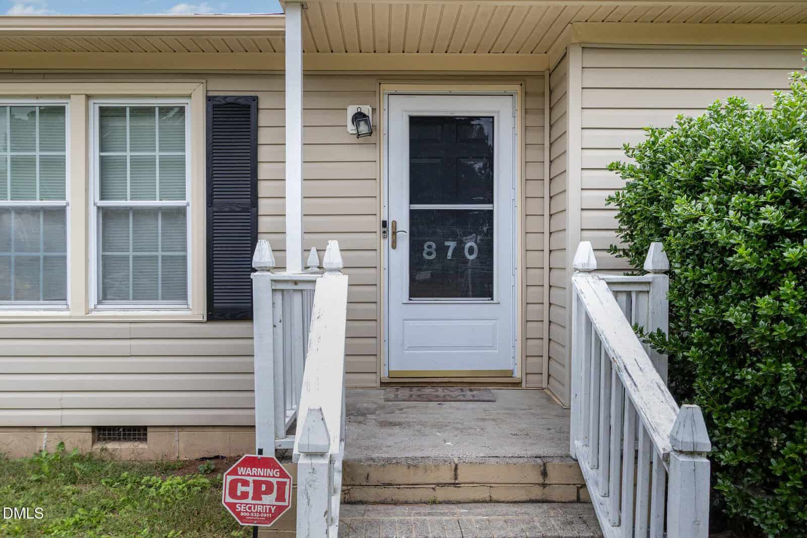 A beige house entrance with white trim, a glass door displaying the number 870, white railing, and a black shutter next to three windows. A "Warning CPI Security" sign is in the grass by the steps.