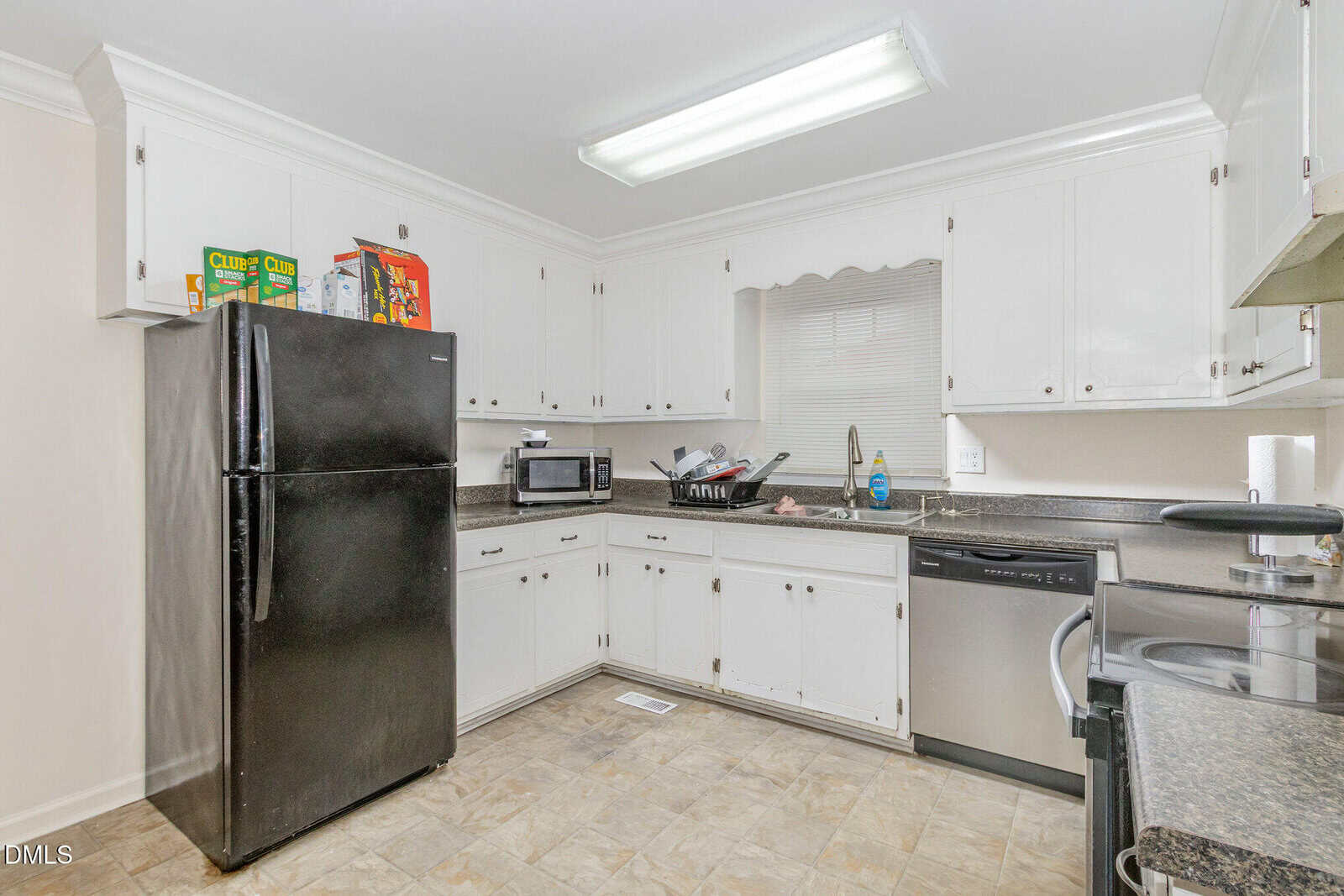 A kitchen with white cabinets, black refrigerator, dishwasher, oven, microwave, and drying dishes by the sink. Cereal boxes are on top of the fridge, and a tiled floor is visible. Bright ceiling light overhead.