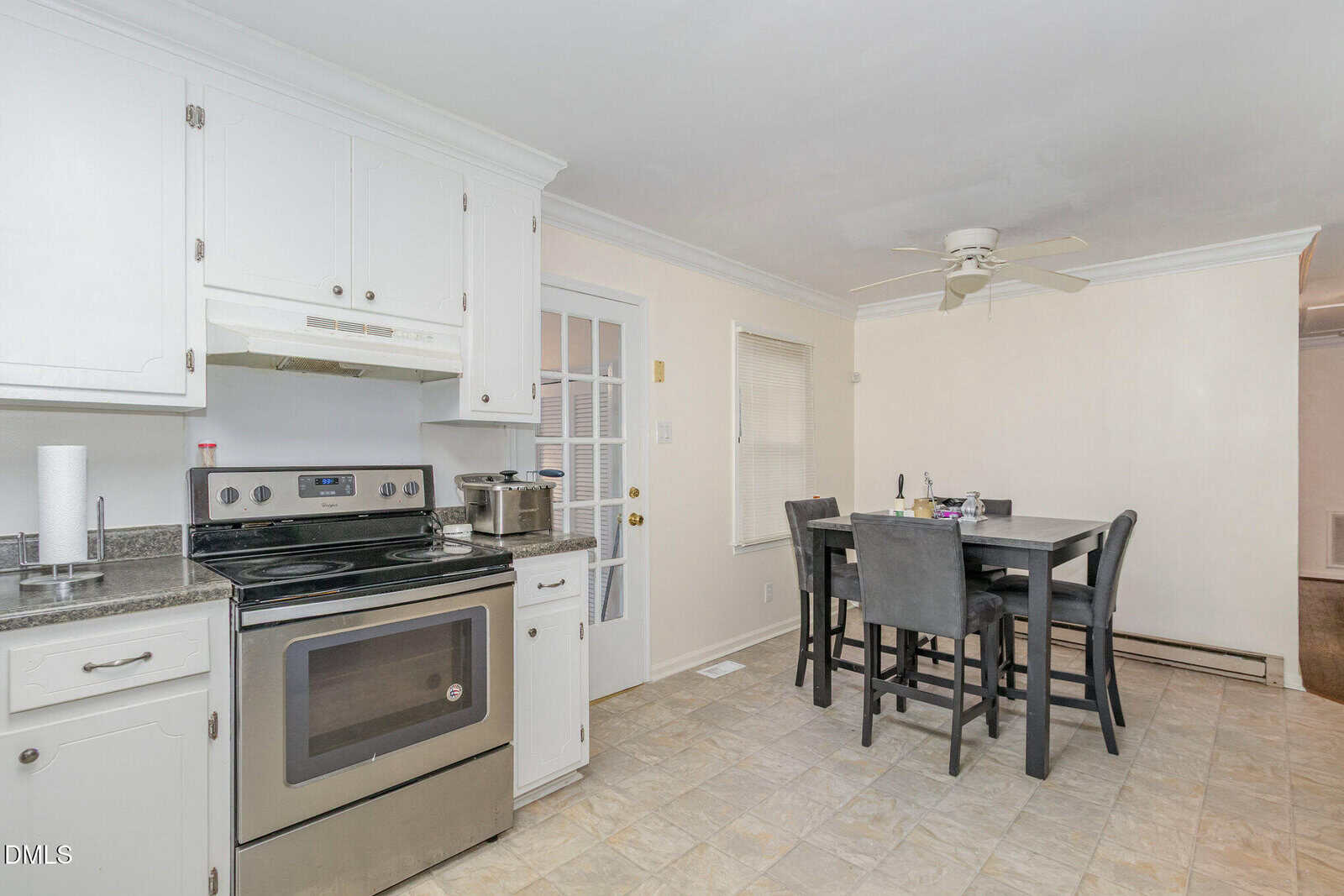 A bright kitchen with white cabinets, stainless steel stove, black countertops, and tile floor. A dining area with a dark table and four chairs is nearby, under a ceiling fan. A door and window let in natural light.
