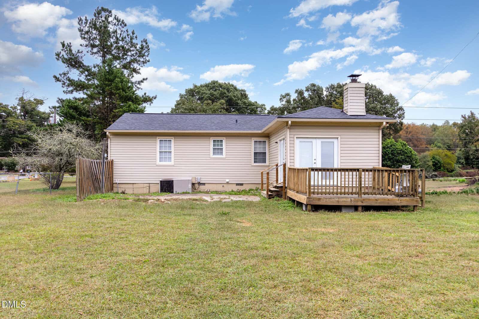 A beige single-story house with a black roof, wooden deck, and small fenced area. The house sits on a large grassy yard with trees and power lines in the background under a partly cloudy sky.