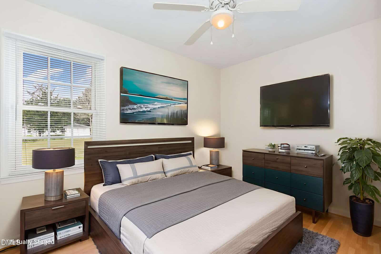 A modern, neatly staged bedroom features a wooden bed with gray bedding, two nightstands with lamps, a dresser with a TV above it, a large wall art piece, a potted plant, and a window letting in natural light.
